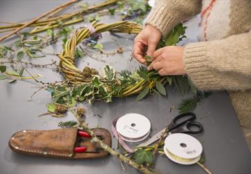 image shows someone making a christmas wreath with natural materials including leaves and holly. There are tools including scissors and ribbon.