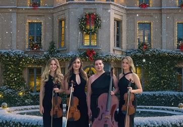 Image shows four women musicians standing outdoors in front of a grand mansion decorated for Christmas. They each hold string instruments — two violin