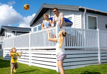 Image shows a caravan at Pevensey with 2 adults on the decking and 2 children playing a ball game on the grass