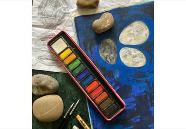 Art workspace scene featuring watercolours and brushes beside a cloudy water cup and several rounded grey stones on a tabletop; warm lighting and shal