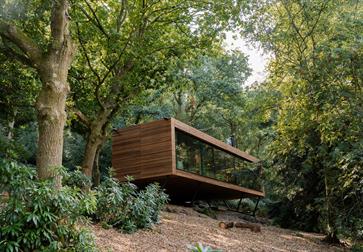 A suspended rectangular wooden lodge with large glass facade in a wooded landscape.