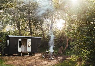 Photograph of a black glamping hut in a woods with smoking fire.