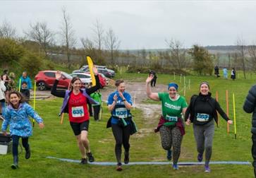 Five runners crossing a finish line on a grassy outdoor course with spectators and cars in the background.