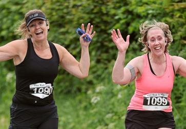 Two runners with race bibs raising their arms while running outdoors on a green, leafy path.