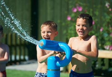 Three young boys dressed in swimming trunks, playing with a fun water sprinkler.