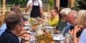 Group of people say at long outdoor picnic table ful lof food. At the head is a man standing, wearing an apron.