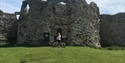 An old castle ruin with grass in the foreground. In front a person poses with their bicycle.