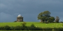 a photograph of a bright green grassy field from a distance, with dark grey moody sky. At the top of the feild is a round domed monument.