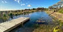 Natural swimming pond at Barnscape Farm with wooden deck, clear water, and surrounding grasses under a bright blue sky.