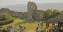 Crowd gathered on grass with stage and performers in front of Hastings Castle ruins in warm evening light.