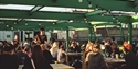 People dining on Bunka Rooftop terrace under green steel beams and string lights in warm evening light.