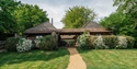 Single-storey timber cottage with pitched roofs, garden path, lawn and white flowering shrubs in a peaceful rural setting.