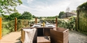 Outdoor dining terrace overlooking countryside, wicker furniture set for a meal with glasses, flowers and rolling green views.