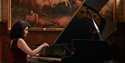 Pianist performing on a grand piano in an ornate hall with a large historic painting in the background.