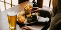 Plate of fish and chips with sauces, a glass of beer and a person reaching for food at a wooden table.