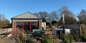 Barnscape Farm cabin with red-framed windows, outdoor seating, garden plants, and a wood-fired hot tub under a clear blue sky.