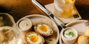 Scotch eggs with dipping sauces on plates, a cocktail with orange slice and a glass of white wine on a wooden table.