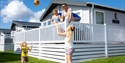 Image shows a caravan at Pevensey with 2 adults on the decking and 2 children playing a ball game on the grass