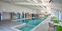 Image shows an indoor pool at the caravan park. The pool is large and rectangle and has a smaller square pool at the start. There are tables and chair