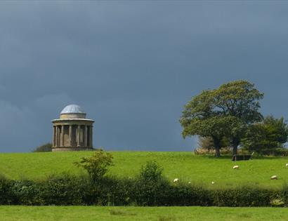 a photograph of a bright green grassy field from a distance, with dark grey moody sky. At the top of the feild is a round domed monument.