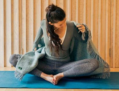 Person sitting cross-legged on a yoga mat, adjusting a fringed blanket in a wooden-panelled room.
