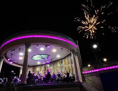 Image shows a band stand with a band playing on it surrounded by purple lights. There is a firework going off in the sky in the background.