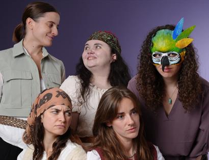 Studio photo of five costumed performers. The rear row features a smiling person in a light utility vest, a person with a patterned headscarf smiling