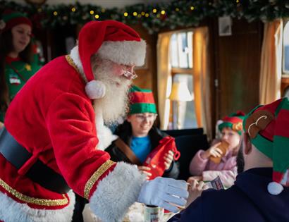 Santa Clause dressed in a traditional red suit with white trim is inside a decorated train carriage on the Kent and East Sussex Railway.