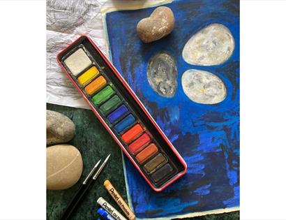 Image show a paint pallet with multiple different colours, some pastel crayons and a sketch drawn on paper
