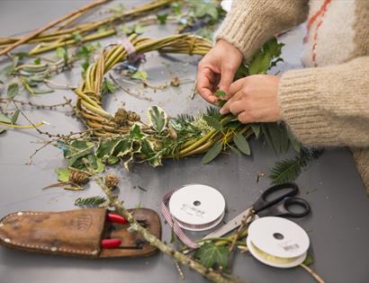 image shows someone making a christmas wreath with natural materials including leaves and holly. There are tools including scissors and ribbon.