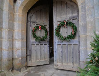 image shows two doors at bodiam castle with christmas wreaths on. In the corner of the image, you can see part of a christmas tree with red baubles on