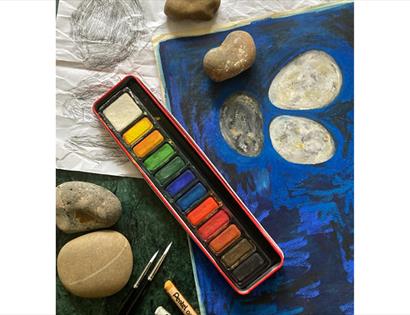 Art workspace scene featuring watercolours and brushes beside a cloudy water cup and several rounded grey stones on a tabletop; warm lighting and shal