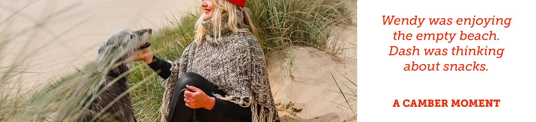 A woman wearing a beanie hat sits on a sandy beach at Camber Sands with her dog.