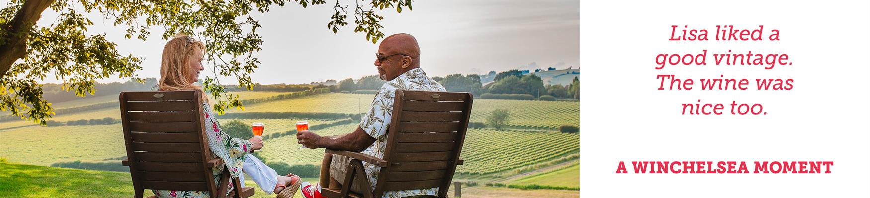With their backs to us, a man and a women sit in low chairs outdoors enjoying a glass of wine with the countryside in the background.