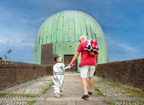 With their backs to the camera, an older man and a young boy walk towards the Herstmonceux Observatory telescope. The boy is dressed like an astronaut and the man has a backpack in the shape of a rocket.