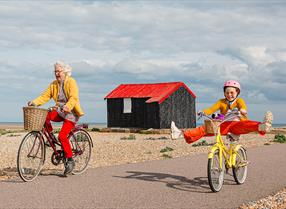 An older lady and a young girl cycle along a wide path at Rye Harbour. A small black hut with a red roof is in the background. They both look excited and the young girl kicks her legs out to each side.