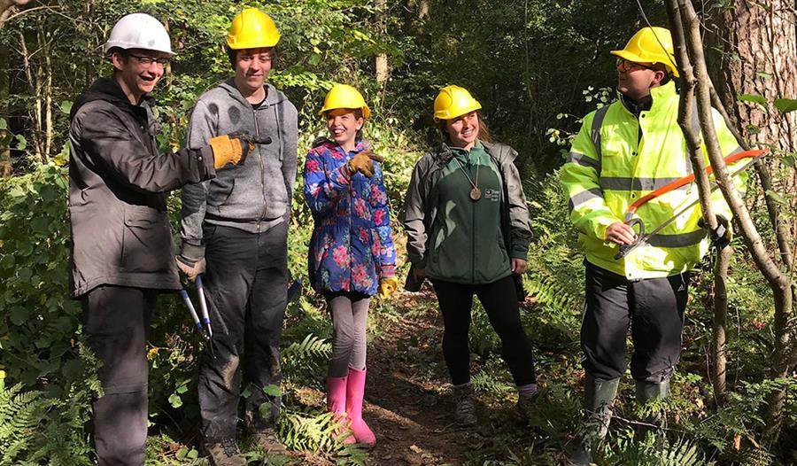a photograph of five people stood in forest with hard hats on. man to right wears high viz jacket whilst others are young adults / teenagers.