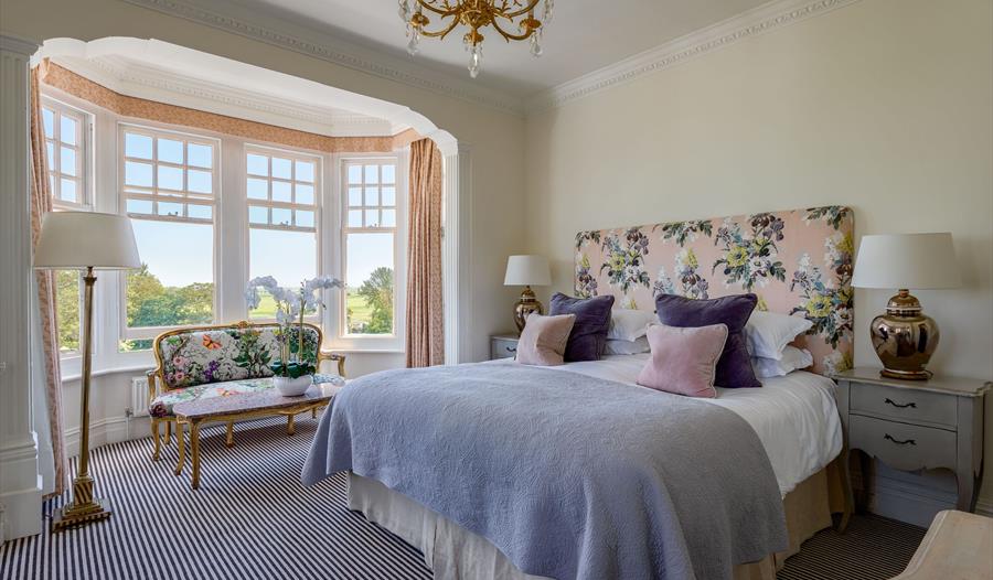 an interior photograph of a hotel bedroom at rye lodge. shows double bed with floral headrest. mauve throw, and bay windows.