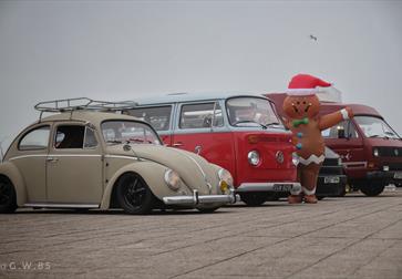 four vintage volkswagens, with beetle in the foreground and three campervans behind. A giant gingerbread character is standing next to them.