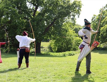 Two men and women extending bows and shooting archery targets on lawn. They are in medieval dress.