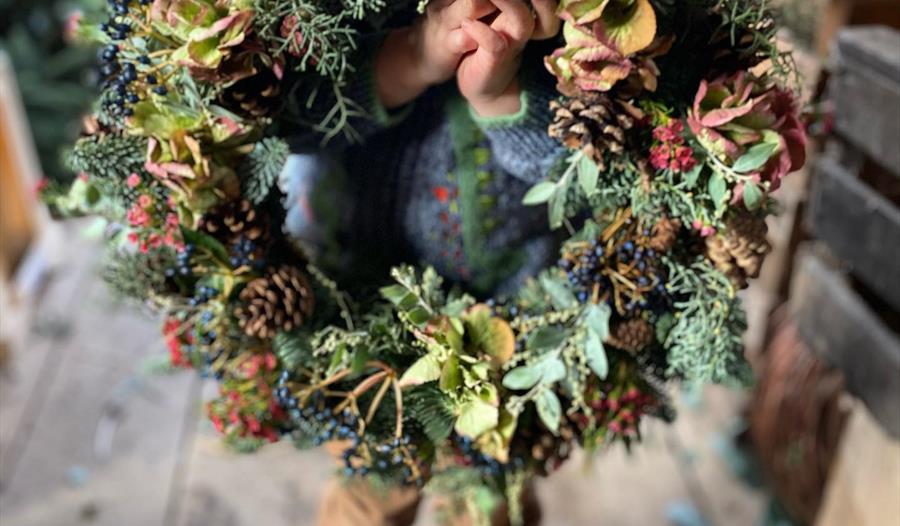 a small child holding a door wreath to the camera, their blue boots visible but their face completely hidden by the wreath.