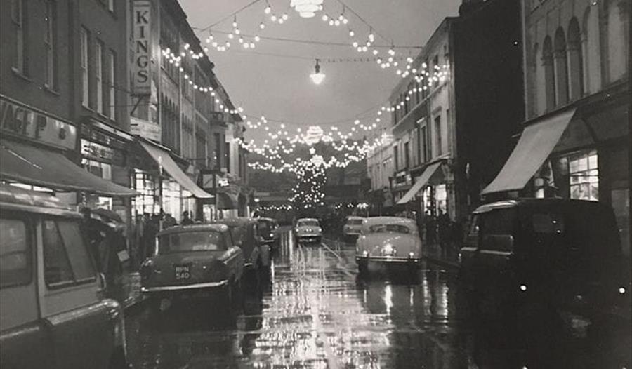 black and white photograph of street scene with 1950s cars and festoon lights reflecting in wet street