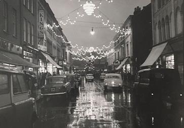 black and white photograph of street scene with 1950s cars and festoon lights reflecting in wet street