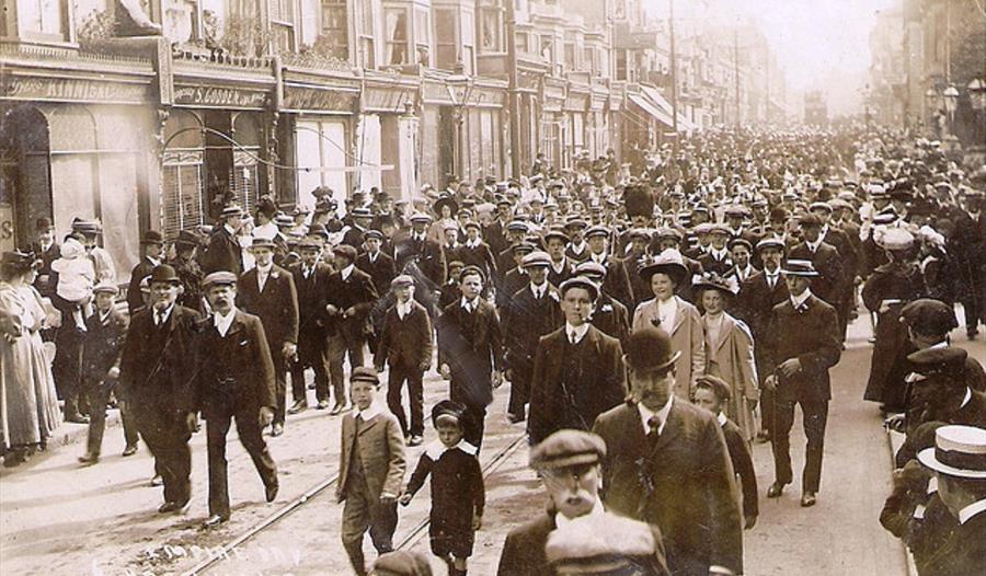 black and white photograph of crowds walking down terraced street, mostly wearing smart clothes and caps.
