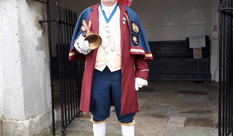 Town crier dressed in traditional dress, burgundy and blue with a tricorn hat with red feathers, holding a large bell.