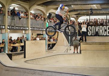 man in mid air on bmx in indoor bowl arena with spectators