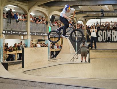 man in mid air on bmx in indoor bowl arena with spectators