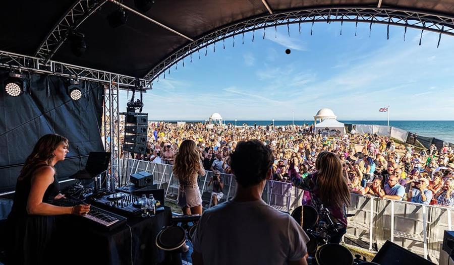 Photograph taken from within outdoor stage, look out at large crowd with the sea in the background.
