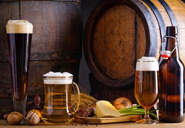 Photograph of three glasses of ales, with wooden barrel in background, surrounded by vegetable and cheese produce.