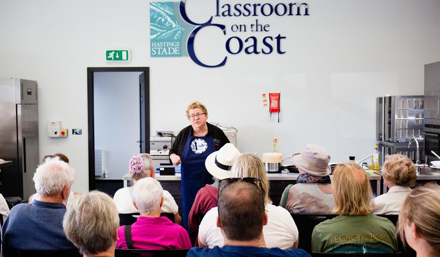 a photograph of a cookery classroom with a teacher, CJ Jackson, in the front, rows of backs of heads.